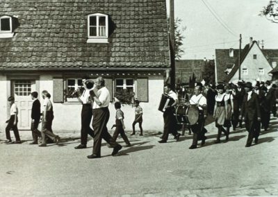Die Kapelle marschiert mit Tenorhorn, Klarinette, Akkordeon und Trommel vor den Schützen am Haus Anselstetter vorbei.